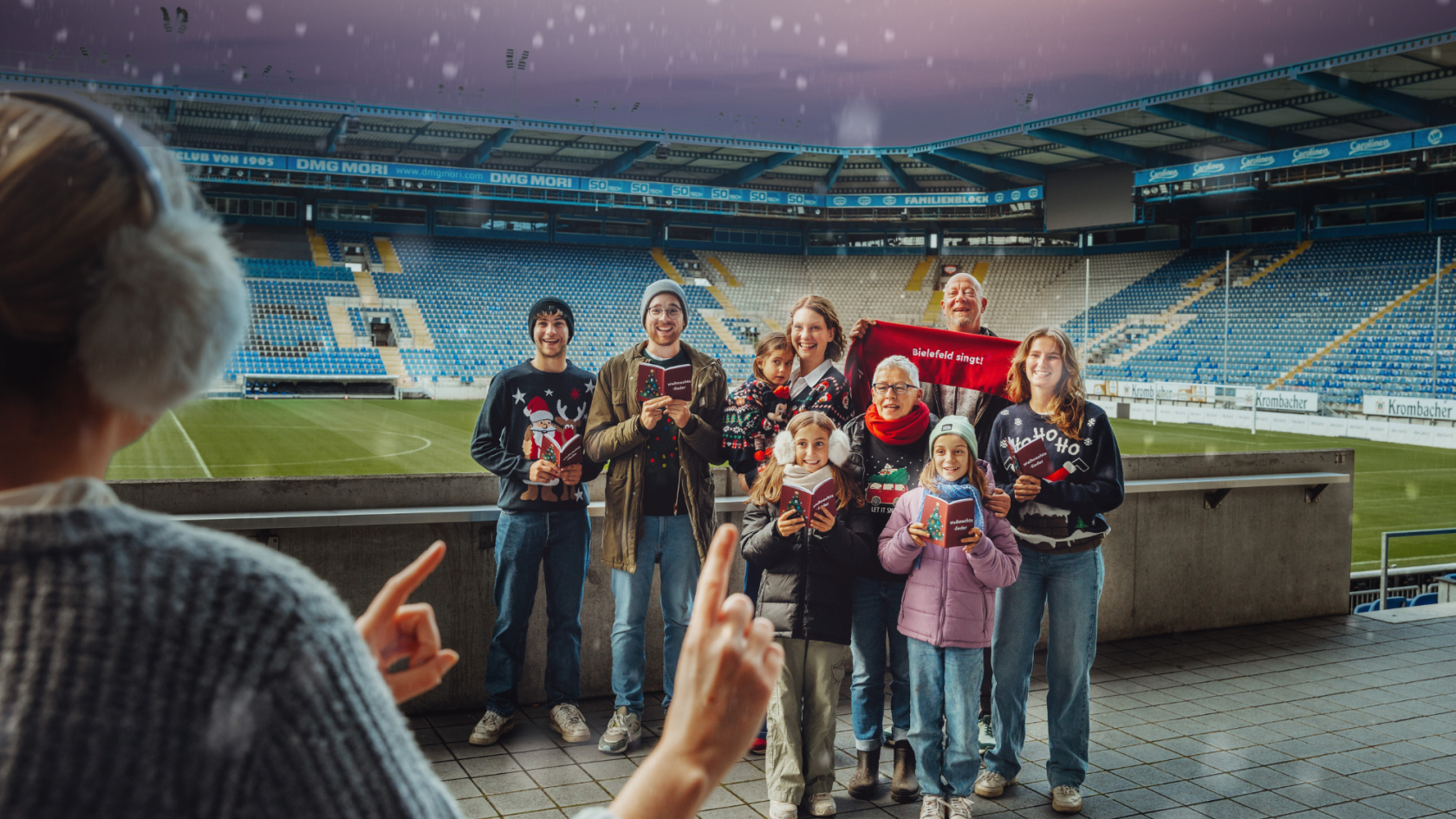 Eine Gruppe von neun fröhlichen Menschen, darunter Kinder und Erwachsene, steht in einem Fußballstadion und singt Weihnachtslieder. Sie tragen bunte Winterkleidung und Weihnachtspullover, einige halten Liedhefte in den Händen. Eine Person im Vordergrund dirigiert die Gruppe. Im Hintergrund sind leere Tribünen und das Spielfeld sichtbar. Ein rotes Banner mit der Aufschrift ‚Bielefeld singt!‘ wird hochgehalten.
