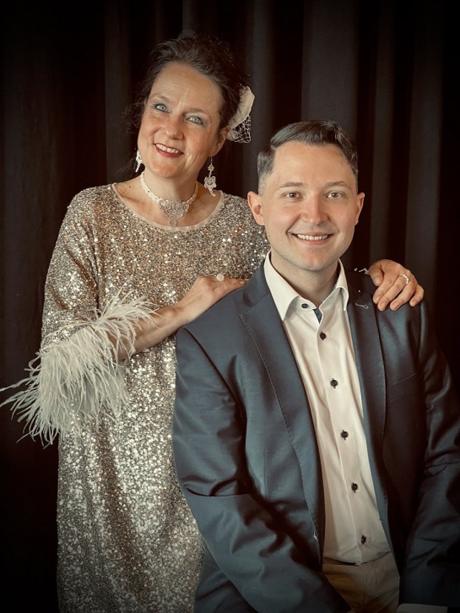 Ein Mann in Sakko sitzend, dahinter eine stehenden Frau im Abendkleid 