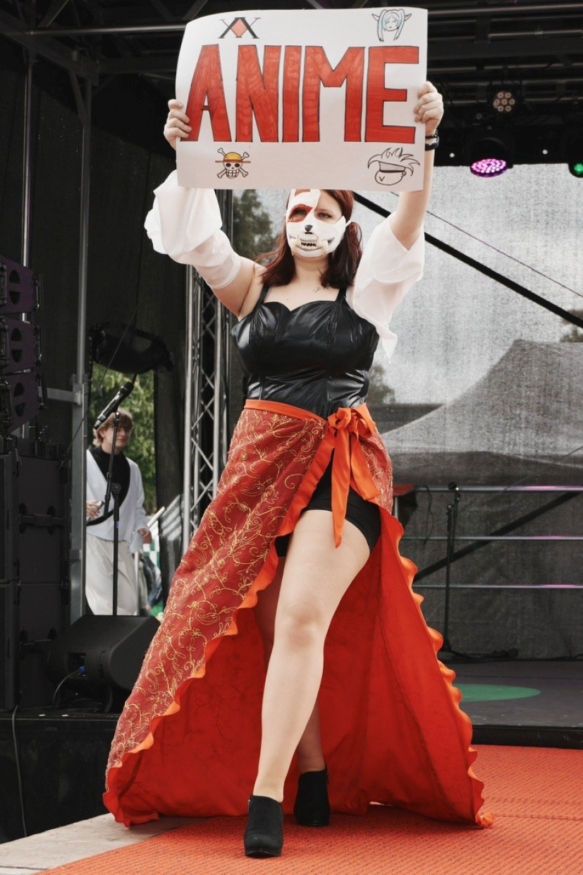 Frau in weißem Crop-Top und rotem Rock auf einer Bühne, hält ein Plakat.