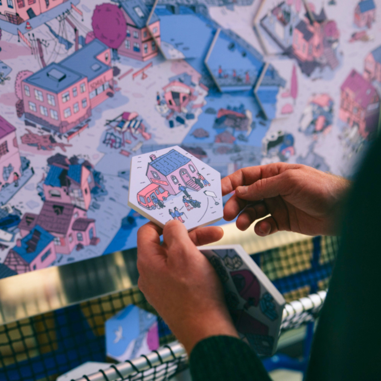 Ein Foto von zwei Händen, welche eine kleine Platte in Form eines Hexagons in der Hand hält. Darauf abegbildet ist ein Haus, ein Auto und ein paar Personen.