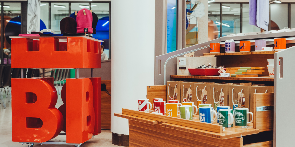 Blick über ein Regal mit Souvenirs, im Hintergrund das Oval der WIssenswerkstadt, am linken BIldrand ein 80x80 großes BIE-Logo