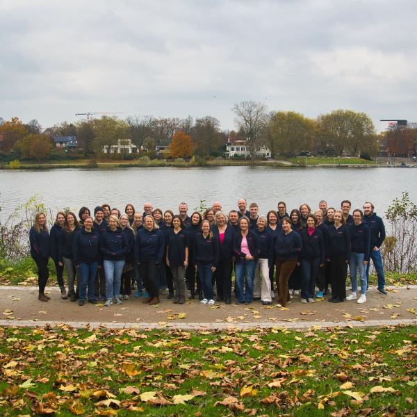 Große Gruppe von etwa 40–50 Menschen steht gemeinsam für ein Gruppenfoto am Ufer eines Flusses. Die Personen tragen überwiegend dunkle, einheitliche Kleidung und schauen in die Kamera. Im Hintergrund sind herbstlich gefärbte Bäume, Häuser und ein bewölkter Himmel zu sehen. Auf dem Boden liegen viele herabgefallene Blätter auf einem Stück Rasen.