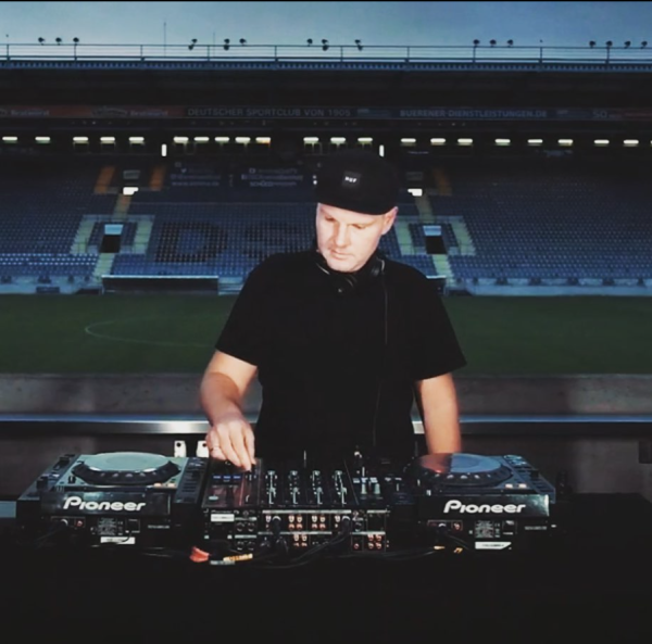 DJ an Mischpult im Stadion, schwarzes Outfit und Hut.