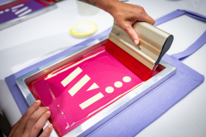 Das Wissenswerkstadt-Logo wird mit einem Siebdruckrahmen auf eine lila Jutetasche gedruckt.