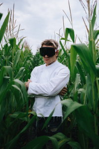 Ein junger Mann (Ski Aggu) mit blonden Haaren steht mit verschränkten Armen in einem Maisfeld. Er trägt eine übergroße schwarze Skibrille, ein weißes, langärmeliges Oberteil und zerrissene Jeans. Der Himmel ist bewölkt, und das hohe Maisfeld umgibt ihn dicht.
