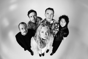 Eine Gruppe Männer und Frauen steht im Kreis und schaut nach oben in die Kamera. Das Foto ist in Schwarz-Weiß gehalten.