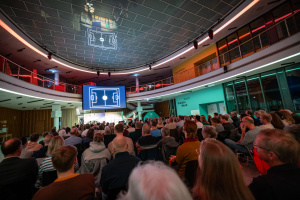 Veranstaltung in einem großen, modern beleuchteten Raum: Publikum schaut auf eine Bühne mit grafischer Spielfeld-Darstellung.