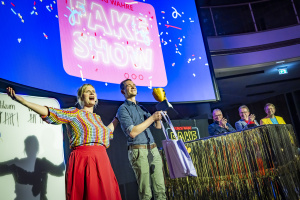 Zwei Moderator:innen auf einer Bühne vor einem großen Bildschirm mit der Aufschrift „Die einzig wahre FAKE SHOW“. Sie jubeln, eine Person hält einen Pokal und eine Geschenktasche. Im Hintergrund applaudieren drei Jury-Mitglieder hinter einem goldglänzenden Pult. Auf der linken Seite ist eine Tafel mit Strichlisten sichtbar. Konfetti fliegt.