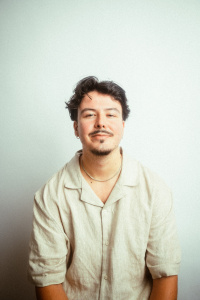 Ein junger Mann mit dunklen, leicht welligen Haaren und einem leichten Bart sitzt entspannt vor einer schlichten, hellen Wand. Er trägt ein beiges, kurzärmeliges Leinenhemd und eine schlichte Silberkette. Sein Gesichtsausdruck ist freundlich mit einem sanften Lächeln. Die Beleuchtung ist weich und verleiht dem Bild eine warme, natürliche Atmosphäre.