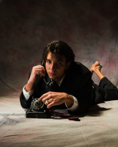 Ein junger Mann mit längeren, unordentlichen Haaren liegt bäuchlings auf dem Boden und hält den Hörer eines klassischen Wählscheibentelefons an sein Ohr, während seine Hand auf dem Telefonapparat ruht. Er trägt einen dunklen Anzug. Das Bild ist in Schwarz-Weiß gehalten und hat eine dramatische Beleuchtung mit starken Schatten.