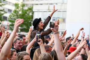 Eine Menschenmenge feiert ausgelassen bei einem Open-Air-Festival. Eine junge Frau mit schwarzem Hut und Netzoberteil sitzt auf den Schultern einer anderen Person, hebt die Arme und lächelt. Viele Festivalbesucher halten ihre Hände in die Luft oder machen Fotos mit ihren Smartphones. Im Hintergrund sind die Universität Bielefeld und grüne Bäume zu sehen.