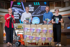 Vier Personen stehen in einem modernen Foyer hinter einer Palette mit Programmheften für die GENIALE 2025. Die Kartons sind mit Wimpelketten und Etiketten dekoriert. Im Hintergrund ist ein großer Bildschirm mit dem Veranstaltungsplakat zu sehen: „GENIALE – KI und wir. Sep.–Nov. 2025“. Alle halten Flyer in der Hand und lächeln in die Kamera.
