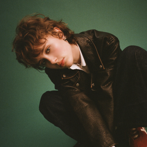 Eine junge Frau mit kurzen, braunen Haaren sitzt mit angezogenen Knien auf dem Boden, Sie trägt eine schwarze Lederjacke und eine schwarze Hose.