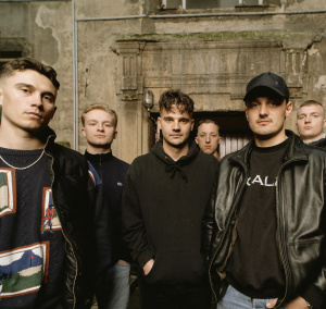 Sechs junge Männer in schwarzer Kleidung stehen in einer Reihe vor einem verwitterten Gebäude. Die Gesichter sind ernst.