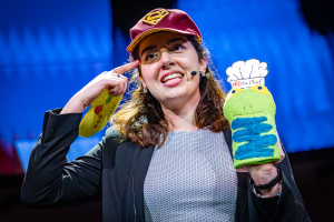 Eine junge Frau hält eine Puppe in der Hand und gestikuliert mit ihrer rechten Hand.